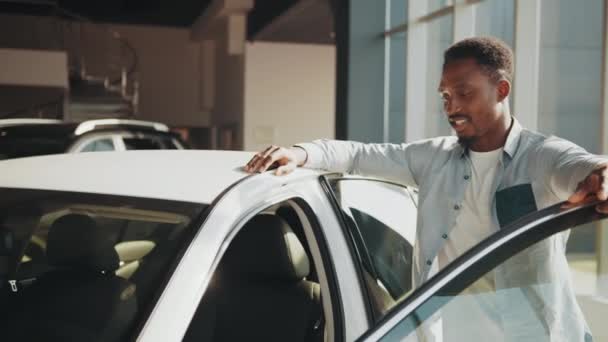 Hombre africano positivo en ropa casual de pie cerca de auto blanco con puerta abierta y examinando el interior. Cliente masculino eligiendo un coche nuevo en la sala de exposición. Concepto de personas y concesionario. Hombre examinando coche nuevo — Vídeos de Stock