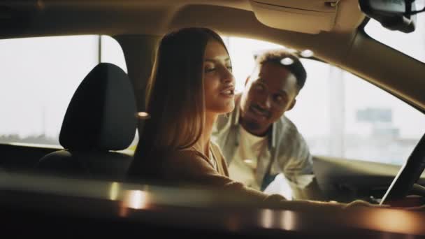 Afro-amerikan delaer yanında durup danışmanlık sağlarken, neşeli beyaz kadın lüks arabanın içinde oturmuş deriyi test ediyor. Otomobil işini kavra. — Stok video