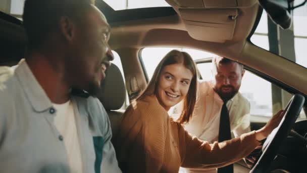 Boldog többnemzetiségű pár ül a modern autóban, miközben az illetékes kereskedésvezetővel beszélgetnek. Új jármű kiválasztása. A drága vásárlás fogalma. Sokszínű pár ül az autóban — Stock videók