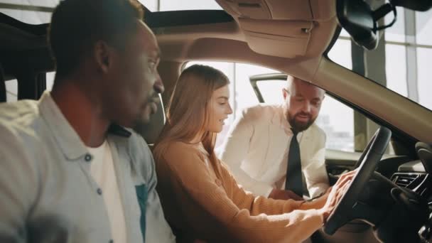 Pareja multiétnica positiva sentada dentro de un coche negro de lujo y hablando con un distribuidor competente en la sala de exposición. Hombre y mujer eligiendo un vehículo nuevo. Auto negocio y concepto de consulta. Multi pareja — Vídeos de Stock