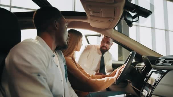 Pozytywny i przyjazny sprzedawca rozmawiający z parą w salonie samochodowym. Afrykanin i biała kobieta siedzą w samochodzie i badają wnętrze skóry. Koncepcja zakupu i transportu. Kupowanie samochodu w salonie — Wideo stockowe