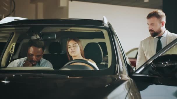 Positivt par sitter inne lyx svart bil och prata med kompetent återförsäljare på showroom. Man och kvinna väljer nytt fordon. Auto företag och samråd koncept. Köpa bil på salongen — Stockvideo