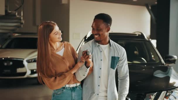 Primer plano de la feliz pareja multicultural posando en el moderno centro de concesionarios con llaves en las manos. Joven hombre y mujer comprando un vehículo nuevo. Satisfacción por la compra costosa. Pareja posando clave — Vídeos de Stock