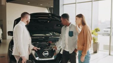 Beyaz tenli satıcı, kapüşonlu lüks bir arabanın yanında duruyor ve kuafördeki müşterilere detaylı inceleme sağlıyor. Çift arabanın yanında satıcıyla duruyor ve satın almadan önce tüm detayları inceliyor..