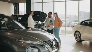 Beyaz tenli satıcı, kapüşonlu lüks bir arabanın yanında duruyor ve kuafördeki müşterilere detaylı inceleme sağlıyor. İnsan kavramı, otomobil işi ve satış. Yeni araç seçiliyor. Araba almak.