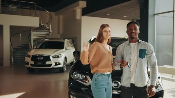 アフリカ人男性と白人女性が車の購入から幸せを表現するために感情的になって喜びました。現代的なサロンに鍵を手に多人種のカップルが立っている。鍵を手にしたカップル — ストック動画