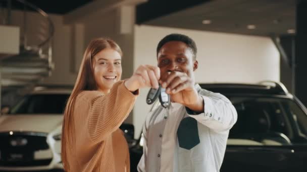 Zbliżenie szczęśliwej wielokulturowej pary pozowanie w nowoczesnym centrum dealerskim z kluczami w rękach. Młody mężczyzna i kobieta kupują nowy pojazd. Satysfakcja z drogich zakupów. — Wideo stockowe