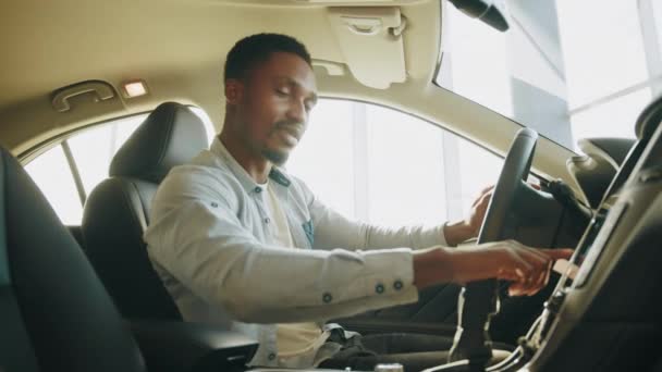 Een man test een nieuwe auto. Zijaanzicht van gefocuste Afrikaanse man die op de bestuurdersstoel zit en dashboard gebruikt voor navigatie. Man op touchscreen op auto multimediapaneel, schakelen van radiostation. — Stockvideo