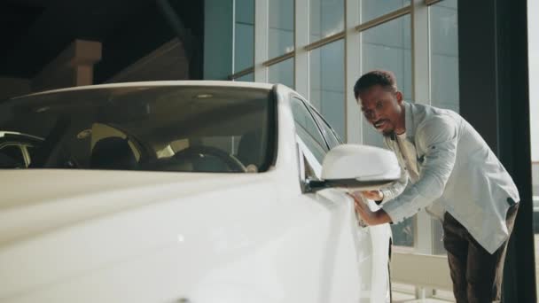 訪問車の販売代理店。アフロ男は新しい車を抱きかかえてドアに触れた。車の販売。サロンで購入した後、幸せなアフロマンは、新しい自動車に触れます。新しい車の概念。男はチェックし、彼の新しい自動車に触れる — ストック動画