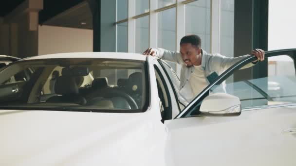 Autokauf. Schöner Mann in lässiger Kleidung fühlt sich vom teuren Kauf im Autosalon befriedigt. Der Käufer öffnet die Autotür beim Händler. Afrikaner begutachtet Neuwagen in modernem Showroom — Stockvideo