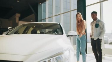 Modern otomobil merkezinde yürürken Afrikalı Amerikalı erkek ve beyaz kadın. Yeni araba seçen, galeride araba alan veya kiralayan mutlu bir milenyum çifti..
