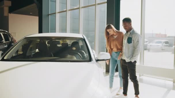 現代の自動車センターを歩いている間にアフリカ系アメリカ人の男性と白人女性。幸せなカップル一緒に新しい車を選択します。車の販売代理店に立って彼らの決定をする美しいカップル — ストック動画