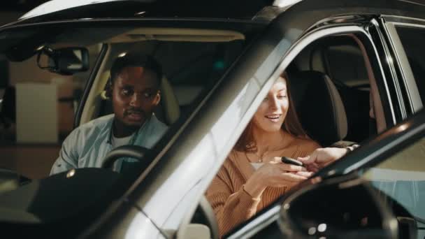 Venditore che dà le chiavi dalla nuova auto. Positiva coppia multietnica seduta all'interno di auto nera di lusso e parlare con rivenditore competente presso lo showroom. Uomo e donna che scelgono il nuovo veicolo. — Video Stock