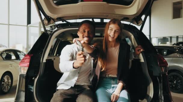 Θετικό πολυφυλετικό ζευγάρι κάθεται σε αγκαλιές στο ανοιχτό πορτ-μπαγκάζ του νέου τους αυτοκινήτου και κρατώντας κλειδιά στα χέρια. Αφρικανός και καυκάσιος αγοράζουν νέο όχημα στο σύγχρονο εκθεσιακό χώρο. Ενώ στο ανοικτό πορτ-μπαγκάζ του αυτοκινήτου — Αρχείο Βίντεο