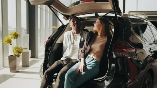 Yeni arabalarının bagajında kucaklaşan pozitif, çok ırklı bir çift. Afrikalı erkek ve beyaz kadın modern galeride yeni bir araç satın alıyorlar. Açık araba bagajında. — Stok video