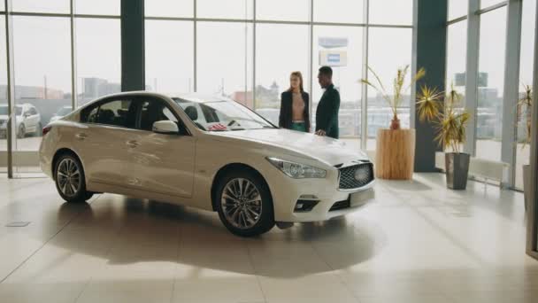 Feliz pareja multi étnica examinando varios auto en la sala de exposición moderna. Hombre africano y mujer caucásica elegir nuevo vehículo juntos. Concepto de venta y compra. Auto en el showroom — Vídeo de stock