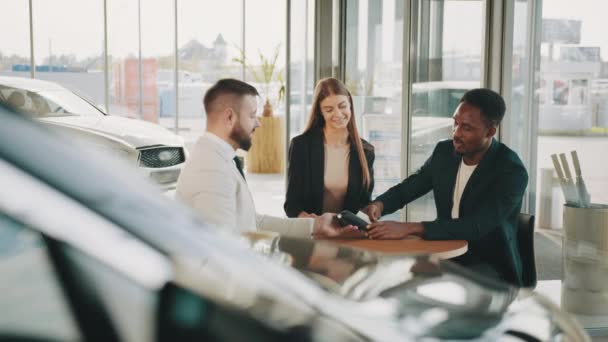 Comprar un coche nuevo. Hombre afroamericano feliz utilizando la tarjeta de crédito para el pago al comprar un coche nuevo. Pareja multiracial sentada con vendedor en salón de automóviles y firmando contrato sobre la compra de coches. — Vídeos de Stock