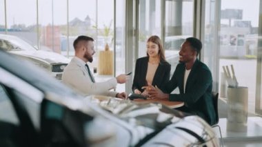 Otomobil galerisinde satıcıyla oturan ve yeni arabalarının anahtarlarını alan mutlu çok kültürlü çift. Gülümseyen adam ve kadın kredi kartıyla ödeme yapıyorlar. Araba galerisinde oturan çift alışveriş yapıyor.