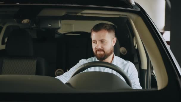 Homme caucasien positif assis sur le siège du conducteur de voiture de luxe, souriant à la caméra et tenant la main sur le volant. Examen du véhicule avant l'achat. Homme examinant le véhicule au salon — Video