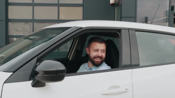 Bělošský vousatý muž si bere bílé luxusní auto na zkušební jízdu ze showroomu. Muž sedí uvnitř moderního auta s červenou mašlí na kapotě. Nové bílé luxusní auto s velkým červeným lukem v moderním salonu — Stock video