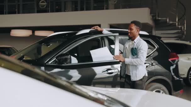 Θετικός Αφρικανός με casual ντύσιμο στέκεται κοντά σε λευκό αυτοκίνητο με ανοιχτή πόρτα και εξετάζει το εσωτερικό. Αρσενικό πελάτη επιλέγοντας νέο αυτοκίνητο στο showroom. Έννοια των ανθρώπων και της αντιπροσωπείας. Άνθρωπος που εξετάζει νέο αυτοκίνητο — Αρχείο Βίντεο