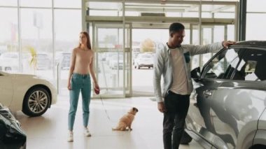 Köpekli mutlu çift yeni bir araç seçiyor. Modern otomobil merkezinde yürürken Afrikalı Amerikalı erkek ve beyaz kadın. Otomobil işi ve danışmanlık konsepti. Çeviri: teşekkür ederim.