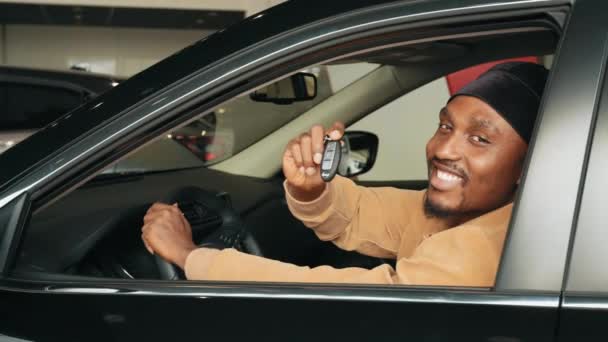 Carro próprio. Homem africano animado mostrando uma chave de carro dentro de seu novo veículo. A visitar a concessionária de carros. alegre afro-americano cara segurando auto chave no automóvel no negócio loja sentado no novo carro — Vídeo de Stock