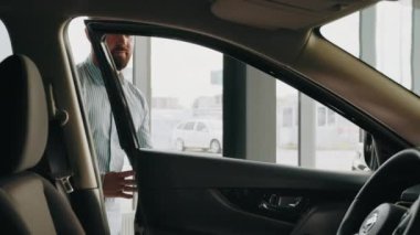 Beyaz adam galeride lüks bir araba açıyor ve ağır çekimde sürücü koltuğunda oturuyor. Adam galeride araç seçiyor. Sakallı genç adam araba galerisinde arabaya dikkatlice bakıyor..