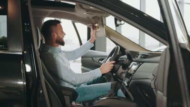 Vrolijke man met baard zittend in een moderne auto met open deur en oprecht lachend op de camera. Gelukkige jongeman koopt nieuwe auto in de moderne showroom. Vrolijke man koopt nieuwe auto in de moderne showroom — Stockvideo