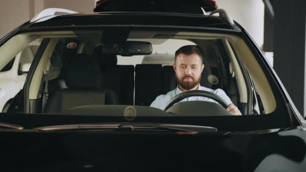Счастливый владелец. Красивый бородатый взрослый мужчина сидит расслабленный в недавно купленной машине и радостно улыбается в окно. Счастливый мужчина сидит в машине и проверяет функцию новой модели в автосалоне. — стоковое видео