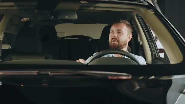 一个积极的高加索人坐在豪华轿车的司机座位上，对着相机微笑，手握方向盘。购车前检查车辆.自己的车。参观现代陈列室时 — 图库视频影像