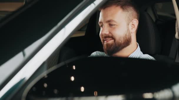 Mann betrachtet Auto in einem Autohaus. Porträt glücklich lächelnder junger Mann Käufer sitzt in seinem neuen Auto aufgeregt bereit für die Reise isoliert vor dem Autohaus viel Büro. Autokauf-Konzept — Stockvideo