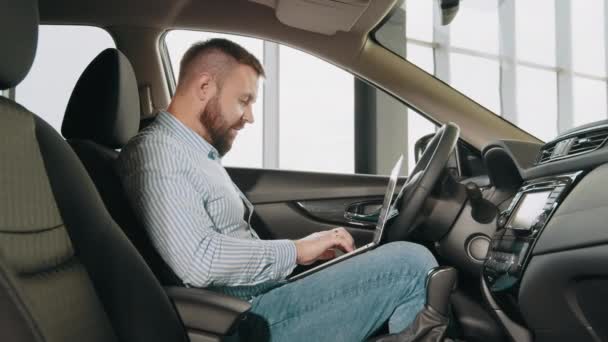 Caucasian bearded man using modern laptop while choosing new car at showroom. Happy man surfing some information in internet while buying new transport. Man using laptop while choosing car at showroom — Stock Video