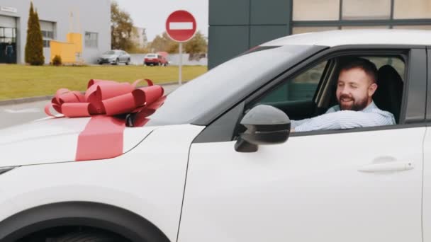 New white luxury car with big red bow riding out of modern auto salon. Expensive purchase of buyer. Caucasian bearded man taking white luxury car for test drive from showroom. — Stock Video