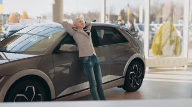 Eko araba satışı konsepti, orta yaşlı, çekici, şık ceketli bir iş kadınının portresi araba galerisinde elektrikli araba seçmesi. Eko araba satışı konsepti. Galeride elektrikli araba.