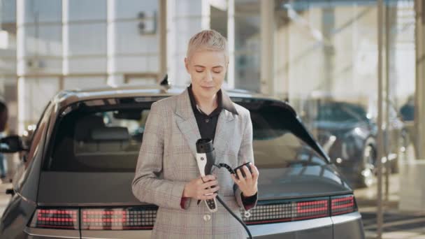 Mujer rubia caucásica de mediana edad sonriente en una estación de carga de coches eléctricos. Hermosa mujer rubia gerente en traje de negocios de pie en el centro de concesionarios de coches eléctricos con cable de carga. — Vídeos de Stock