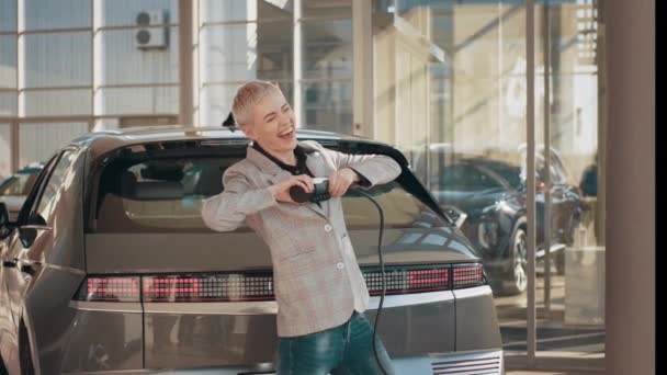 Eco concepto de coche eléctrico. Mujer rubia juguetona en una estación de carga de coches eléctricos. Hermosa mujer rubia gerente en traje de negocios bailando en el centro de concesionarios de coches eléctricos con cable de carga. — Vídeos de Stock