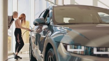 İki mutlu genç kadın, otoparktaki şarj istasyonunda elektrikli arabaları şarj olana kadar birbirleriyle konuşuyorlar. Çevre dostu ulaşım konsepti. Eko elektrikli araba.