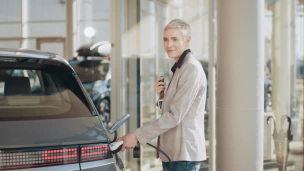 Una mujer cerca de su coche eléctrico, lo desconecta de la carga en el estacionamiento. Una mujer de negocios completa la carga y desconecta el cable del coche eléctrico mientras se carga en el centro de negocios — Vídeos de Stock