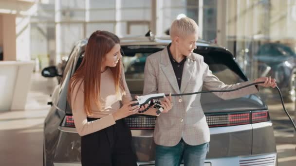 Młoda uśmiechnięta rodzina lgbt kupuje pierwszy elektryczny samochód w salonie. Ochrona środowiska. Atrakcyjne Dwie kobiety trzymające kabel z elektrycznego samochodu i elektrycznej wtyczki ładowania samochodu. — Wideo stockowe
