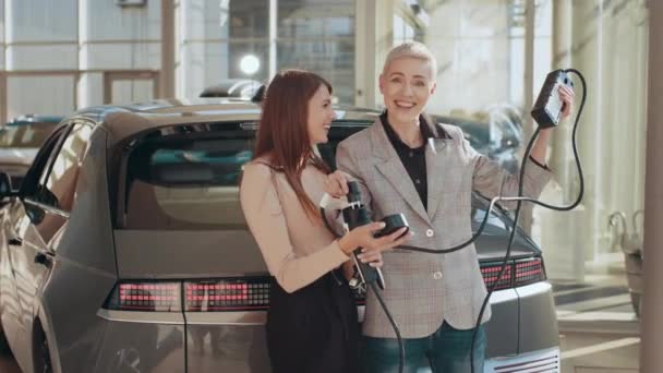 Młoda uśmiechnięta rodzina lgbt kupuje pierwszy elektryczny samochód w salonie. Ochrona środowiska. Atrakcyjne Dwie kobiety lgbt trzymające kabel z elektrycznego samochodu i elektrycznej wtyczki ładowania samochodu. — Wideo stockowe