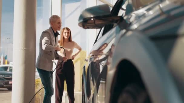 Dwie piękne kobiety w garniturze biznesowym stojące w centrum sprzedaży samochodów elektrycznych trzymające kabel do ładowania samochodów elektrycznych. Kobieta wyciąga złącze z elektrycznego samochodu. Koncepcja samochodu elektrycznego. — Wideo stockowe