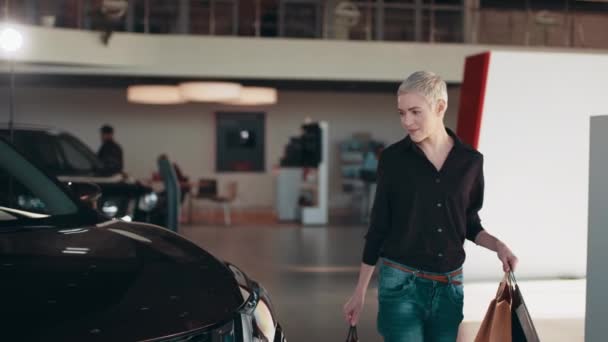 Female buyer client choose auto want to buy new automobile in car showroom. Blond short hair woman in black shirt stands in car salon near vehicle with package in hands. Sales shopping concept. — Vídeos de Stock