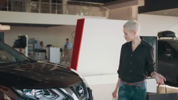 Blond short hair woman in black shirt stands in car salon near vehicle with package in hands. Sales shopping concept. Female buyer client choose auto want to buy new automobile in car showroom — Video