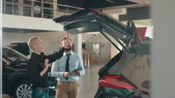 Blond female customer with salesman inspects car at car dealership. The man shows the woman how to close the electric trunk in the crossover. The lady and the salesman inspect the trunk of the machine — Vídeos de Stock