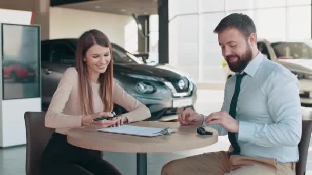 A beautiful girl signs documents to buy a car and receives the keys from the seller. A happy client enters into a successful deal in a modern dealership. The car dealer gives the keys to the new car — Vídeos de Stock