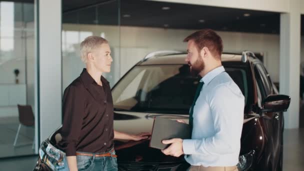 Friendly male car seller with blond female customer in car salon, holding tablet and talking about specifications and performances of automobile. Customers looking at cars in showroom. Cool design. — Vídeos de Stock