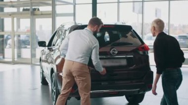 Galeride arabayı inceleyen bir satıcıyla sarışın, güzel bir bayan müşterinin dikiz görüntüsü. Adam, kadına çapraz ateşte otomatik bagajın nasıl açılacağını gösteriyor. Satış merkezinden araba almak..