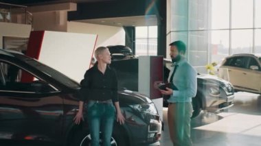 Yakışıklı adam, yetenekli araba satıcısı müşterileriyle çalışmak için dijital tablet kullanıyor. Orta yaşlı, zayıf, sarışın bir iş kadını, satış merkezinden araba alırken müdürle konuşuyor..