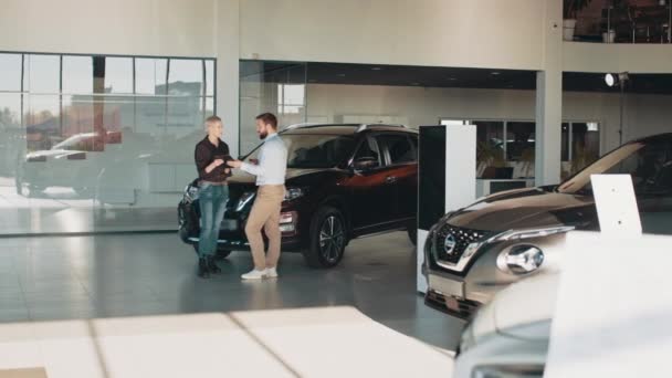 Serious male car seller walking around car salon with blond short hair business woman who wants to buy a car and talking about specifications of cars. Concept of people and transport. — Vídeos de Stock